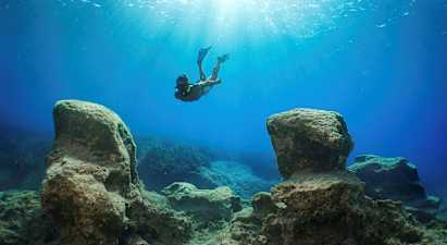 En snorklare simmar under vattnet mellan stora klippor med solljus som filtreras ner från ytan. (AI-genererad text)