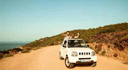 En vit bil parkerad på en grusväg vid sidan av en kustlinje med skogklädda kullar. (AI-genererad text)