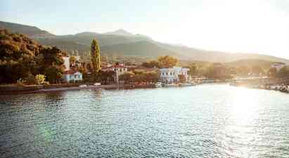 En liten by ligger vid en vattenkant med hus och träd omgivna av berg. Solen skiner över området. (AI-genererad text)