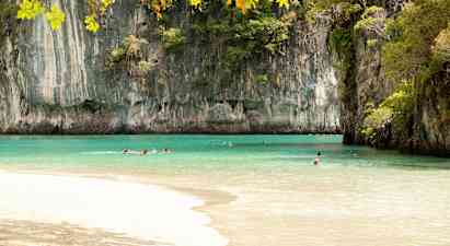 Bilden visar en strand med klart turkost vatten omgiven av höga klippformationer täckta med växtlighet. Flera personer simmar i vattnet under en klar himmel. (AI-genererad text)