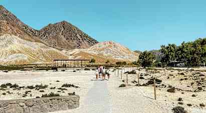 En torr landskapsvy med personer som går på en stig som leder mot några små byggnader. Berget syns i bakgrunden under en klarblå himmel. (AI-genererad text)