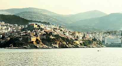 Bild som visar en kuststad med bebyggelse på en höjd nära havet, med båtar på vattnet och gröna kullar i bakgrunden. (AI-genererad text)