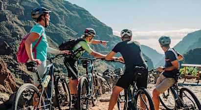 En grupp människor med mountainbikes tittar på en naturskön utsikt mellan berg med grönskande dalar. (AI-genererad text)