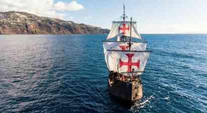 Ett segelfartyg med stora vita segel dekorerade med röda kors navigerar över en blå ocean i närheten av en kuperad kust. (AI-genererad text)