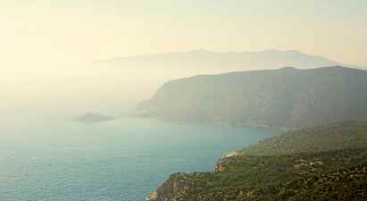 En kustlinje med branta klippor och gröna skogar som sluttar ner mot havet. Ett dis täcker landskapet. (AI-genererad text)