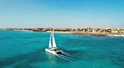 En katamaran med vita segel färdas över turkost vatten nära en kust med hus i olika färger. (AI-genererad text)