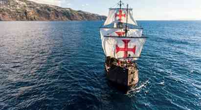 Ett segelfartyg med stora vita segel dekorerade med röda kors navigerar över en blå ocean i närheten av en kuperad kust. (AI-genererad text)