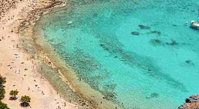 Strand med gyllene sand och klart turkost vatten. Många människor njuter av solen och simmar. En segelbåt ligger för ankar längre ut. (AI-genererad text)