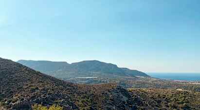 En vy över ett bergigt landskap med varierande växtlighet och havet i bakgrunden under en klarblå himmel. (AI-genererad text)
