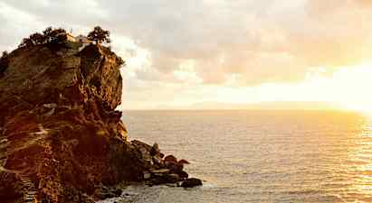 En klippa vid kusten med en byggnad och träd på toppen. Havet syns under en solnedgång. (AI-genererad text)