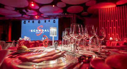 En bordsdukning med vinglas i ett rum belyst med röda lampor. Bakgrund med en stor skärm med texten 'Scandal' och en scen. (AI-genererad text)