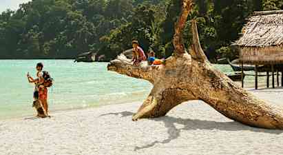 Två barn sitter på en stor trästock på en sandstrand vid havet, omgiven av grönskande skog och träbyggnader. (AI-genererad text)