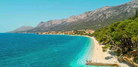 Vy över en klapperstensstrand med turkost vatten och grönskande träd. Berg och en by syns i bakgrunden. (AI-genererad text)