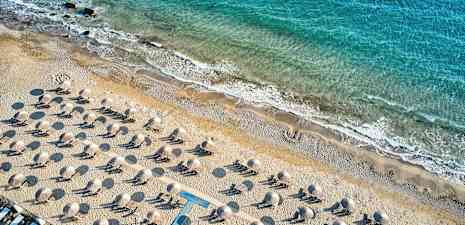 Flygfoto av en sandstrand med solstolar och parasoller, med kustlinjen och blått hav i bakgrunden. (AI-genererad text)