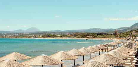 Strand med parasoll av halm vid havet och en bergig bakgrund. (AI-genererad text)