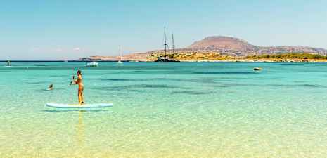 Person som står på en paddleboard i klart blått vatten med en bergig kustlinje i bakgrunden. (AI-genererad text)