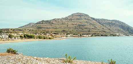 En kust med stenstrand och klart vatten, i bakgrunden syns hus och en hög kulle. (AI-genererad text)