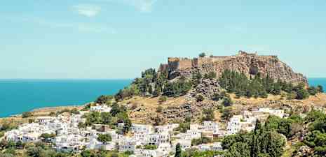 Utsikt över en kulle med en historisk fästning omgiven av gröna träd och vita byggnader nära havet. (AI-genererad text)