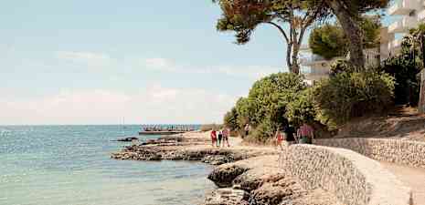 Stenstrand med slingrande gångväg intill havet, omgivet av träd och byggnader. (AI-genererad text)