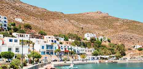 En kuststad med vita och färgglada byggnader, små båtar vid en stenig kustlinje och torra berg i bakgrunden. (AI-genererad text)