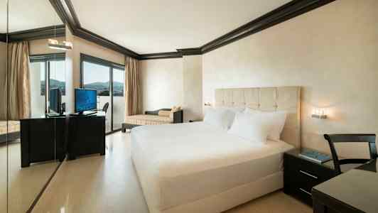 Hotelrum med dubbelsäng, skrivbord med stol och TV samt en stor fönsterdörr. (AI-genererad text)