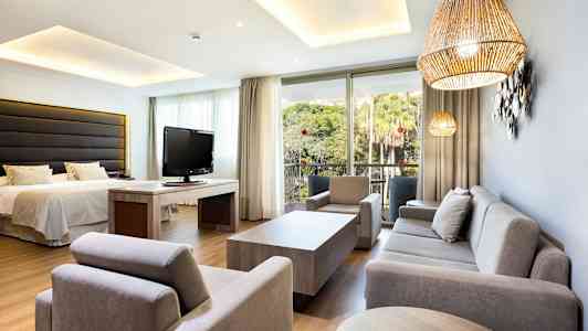 Ett hotellrum, dekorativt möblerat med soffa, fåtöljer, säng och tv, samt utsikt mot en grönskande trädgård. (AI-genererad text)