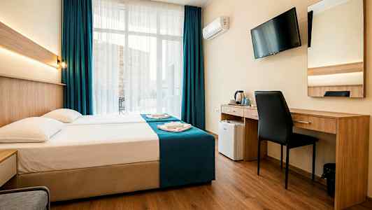 Ett hotellrum med en dubbelsäng, skrivbord, stol, väggmonterad TV och stora fönster med mörkblå gardiner. (AI-genererad text)