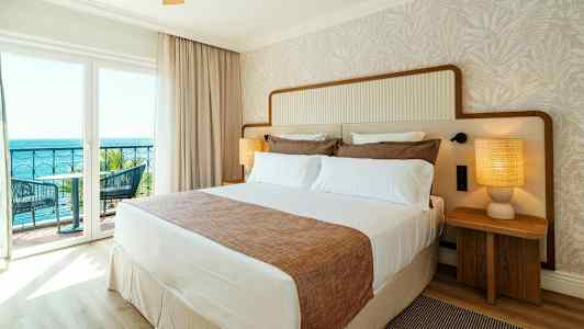 Ett hotellrum med en stor säng, två sängbord med lampor och en balkong med utsikt mot havet. (AI-genererad text)