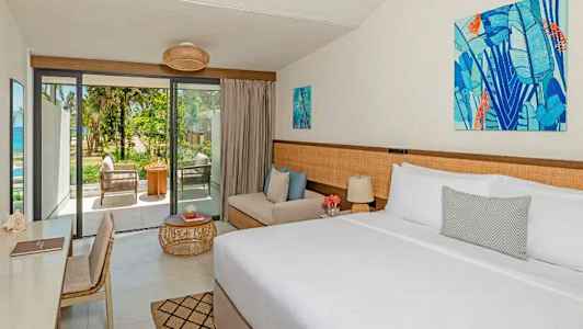 Ett ljust inrett hotellrum med en dubbelsäng, soffa och ett skrivbord. Ett stort glasparti leder ut till en terrass med utsikt mot grönska och en strand. (AI-genererad text)