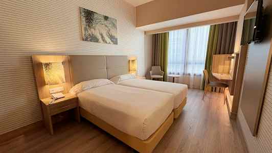 Ett hotellrum med två enkelsängar, nattduksbord, en arbetsyta och en fåtölj bredvid ett fönster. (AI-genererad text)