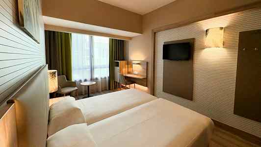 Ett hotellrum med två enkelsängar, en arbetsplats, en stol och ett bord nära ett stort fönster. (AI-genererad text)