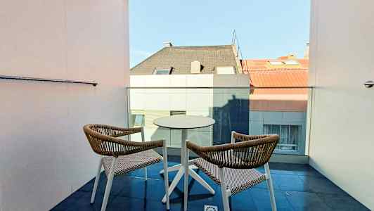 Två stolar och ett bord på en balkong med utsikt mot hustak. (AI-genererad text)