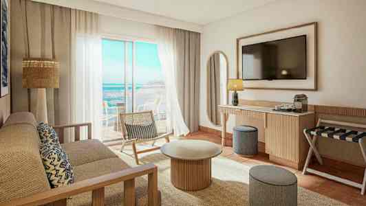 Ett ljust och modernt hotellrum med utsikt över havet. Det finns en soffa, ett soffbord, en stol och en TV på väggen. (AI-genererad text)