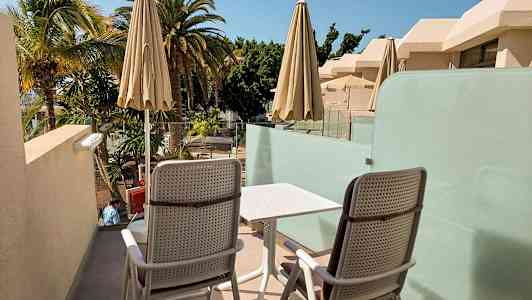 Två stolar och ett bord på en terrass med utsikt över palmer och byggnader. (AI-genererad text)