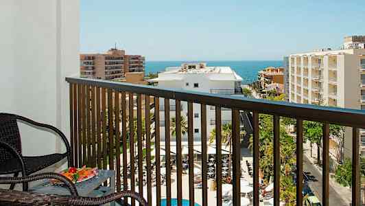 Utsikt från en balkong med möbler över en pool, byggnader och havet i bakgrunden. (AI-genererad text)