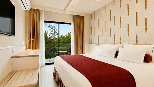 Ett hotellrum med en stor säng täckt av ett rött överkast och vita kuddar, ett bord och två stolar utomhus på balkongen. (AI-genererad text)