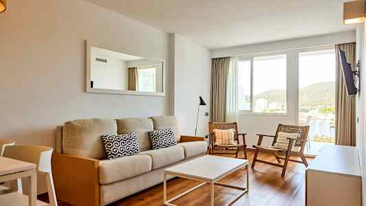 Ett vardagsrum med en beige soffa, två stolar och ett vitt soffbord. Det finns ett fönster med utsikt mot grönska. (AI-genererad text)