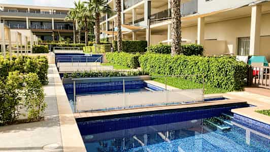 En bild av hotellområdet med flera pooler, gröna buskar och palmträd nära byggnaden. (AI-genererad text)