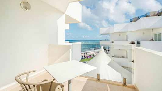 En balkong med bord och stolar, med utsikt över havet och vita byggnader. (AI-genererad text)