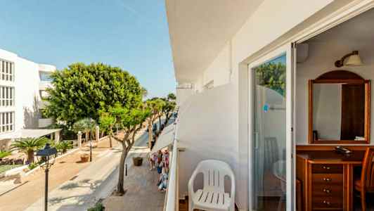 Utsikt från en balkong med en vit plaststol. Gatan nedanför har träd och byggnader. (AI-genererad text)