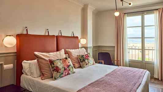 Ett hotellrum med en säng, dekorativa kuddar och ett fönster som visar utsikt. (AI-genererad text)