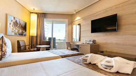 Ett hotellrum med två enkelsängar, sittmöbler vid ett runt bord, samt en skrivbordshörna med en väggmonterad TV ovanför. (AI-genererad text)