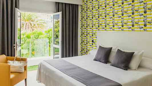 Ett hotellrum med en dubbelsäng och färgstarkt mönstrat tapet. Balkongdörrarna står öppna och visar en grönskande utsikt. (AI-genererad text)