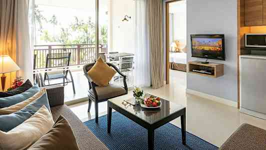 Modernt inrett vardagsrum med soffa, fåtölj, glasbord med frukt, TV och köksdel, samt balkong med utsikt mot palmer. (AI-genererad text)