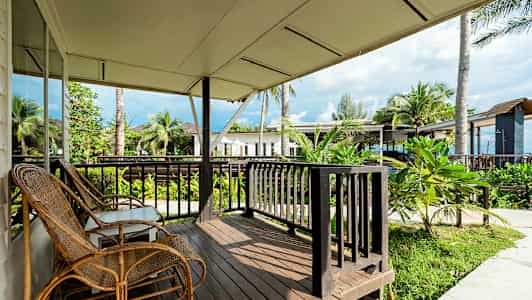 Veranda med trätrall, rottingstolar och utsikt över grönska samt byggnader och palmer i närheten. (AI-genererad text)