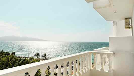 Utsikt från en balkong mot havet och en klar himmel, med palmer och trädkronor längst ner. (AI-genererad text)