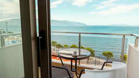 Bild av en balkong med glasräcke, ett bord och två stolar. Utsikten visar havet och en strand med träd. (AI-genererad text)