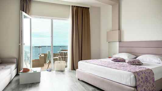 Ett hotellrum med en stor säng, glasdörrar till en balkong och utsikt över havet. (AI-genererad text)