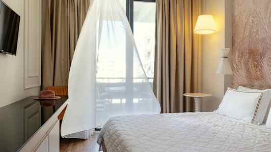 Ett hotellrum med en säng, TV, bord och stol, samt ett fönster med vita gardiner. (AI-genererad text)