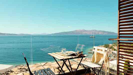 Utsikt från en balkong mot havet och en strand med parasoller. På balkongen finns ett litet bord och stol. (AI-genererad text)